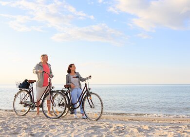 Ein Paar steht mit Fahrrädern am Sandstrand einer Küste mit blauem Wasser und hellem Himmel | © GettyImages.com/FredFroese
