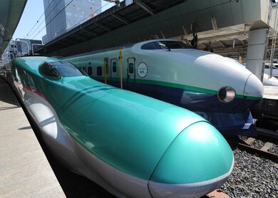 Zwei Shinkansen-Züge stehen nebeneinander auf einem japanischen Bahnhof mit modernen Hochhäusern im Hintergrund | © J-Navigator Foto-Archiv