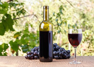 Rotweinflasche mit einem Glas und roten Weintrauben auf einem Tisch im Hintergrund grünes Weinlaub | © GettyImages.com/Hetman Bohdan