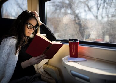 Entspannte Zugreise - Frau sitzt am Fensterplatz im Zug und genießt das Buch und den heißen Kaffee auf dem Tisch vor sich | © Gettyimages.com/DusanManic