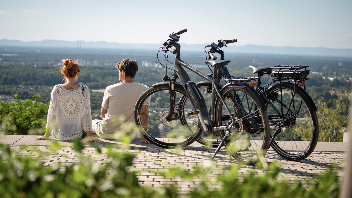 Ein junges Paar sitzt oberhalb eines Berges und genießt die Aussciht auf die Stadt Karslruhe, neben ihnen stehen ihre geparkten Fahrräder | © KTG Karlsruhe Tourismus GmbH/BFC