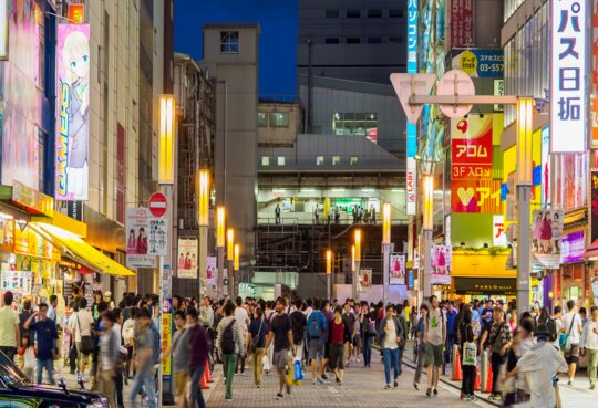 beliebte Strasse in einem Einkaufsviertel bei Nacht, beleuchtet von zahlreichen Leuchtreklamen und gefuellt mit vielen Menschen. | © Gettyimages/kawamura_lucy