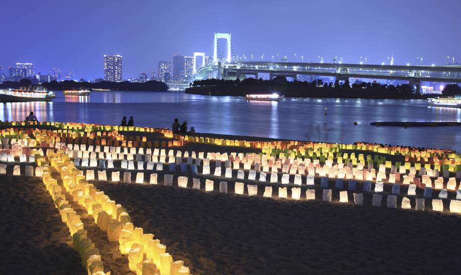Abendstimmung am Meer mit leuchtenden Laternen auf dem Sand und einer beleuchteten Bruecke und Skyline im Hintergrund. | © gettyimages.com/gyro