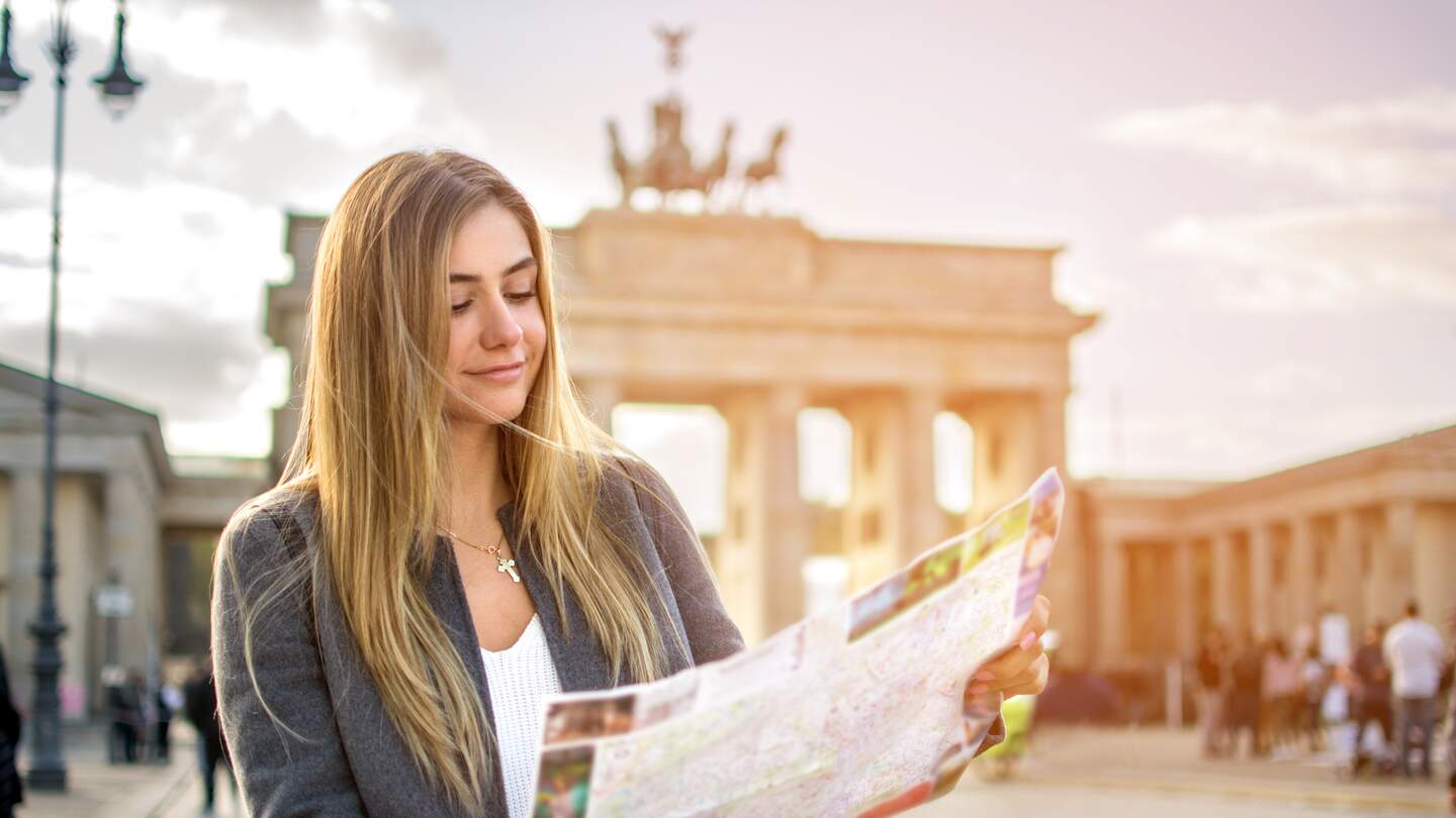 Eine junge Frau mit einer Stadtkarte steht vor dem Brandenburger Tor. Die Sonne scheint. | © Gettyimages.com/Bojan89