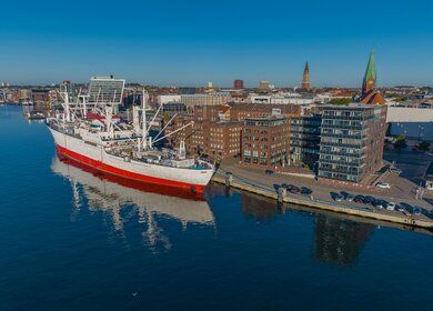 Luftaufnahme des größten Museumsfrachtschiffs der Welt, das im Kieler Hafen vor Anker liegt. Ein weißes und rotes Schiff im Hafen von Kiel. | © Gettyimages.com/frederick doerschem