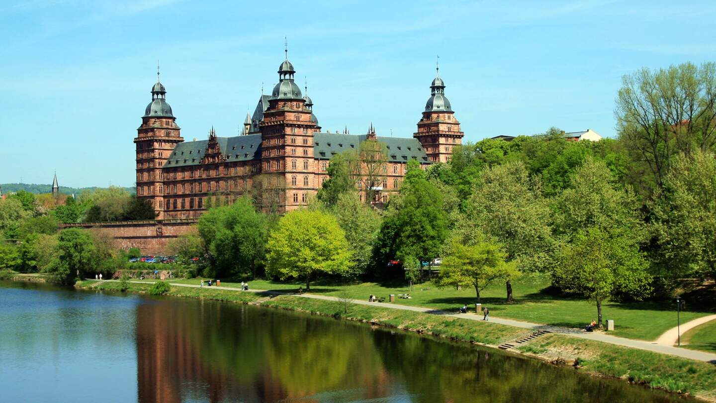 Blick auf Aschaffenburger Schloss Johannisburg mit gruener Wiese und Baeumen angrenzuend zum Main  | © GettyImages.com/wangyangcn