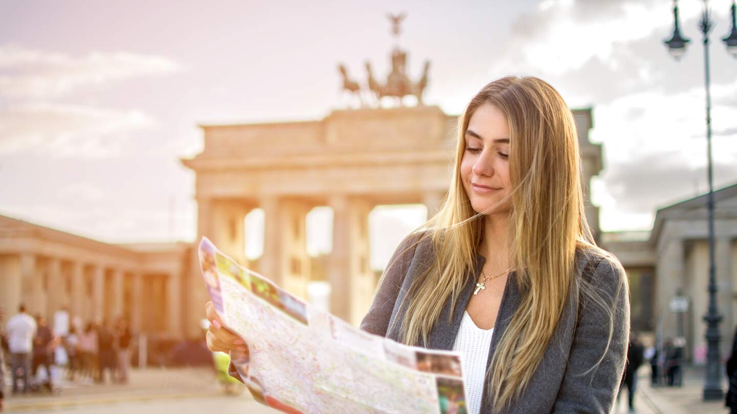 Eine junge Frau mit einer Stadtkarte steht vor dem Brandenburger Tor. Die Sonne scheint. | © Gettyimages.com/Bojan89