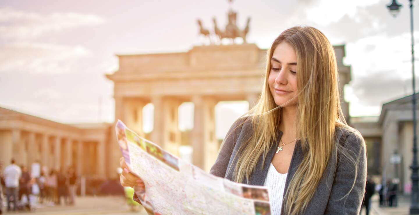 Eine junge Frau mit einer Stadtkarte steht vor dem Brandenburger Tor. Die Sonne scheint. | © Gettyimages.com/Bojan89