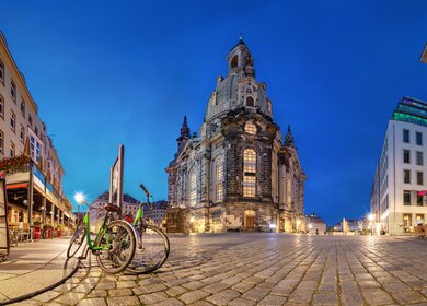 Rückseite der abendlich beleuchteten Frauenkirche Dresden mit Straßencafé und auf gepflastertem Platz geparkten Fahrrädern  | © Gettyimages.com/bbsferrari