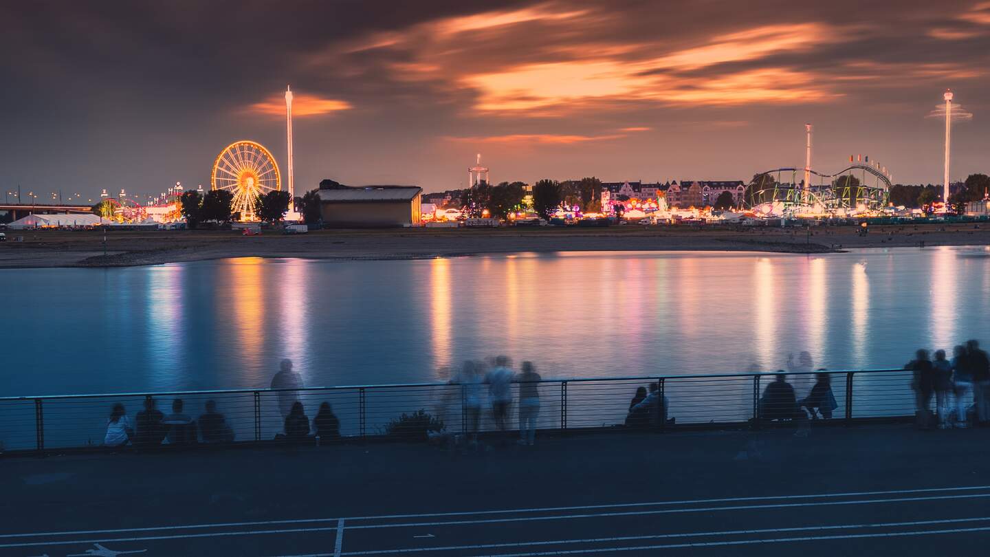 Abendansicht von Messefest und Rheinkirmes bei einem traditionellen Fest am Rheinufer. Menschen, die Riesenrad und andere beleuchtete Attraktionen beobachten | © Gettyimages.com/franticc00