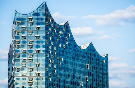 Nahaufnahme der markanten Silhouette der Glasplatte der Hamburger Elbphilharmonie, die ihr glaenzendes und futuristisches Wellendesign vor blauem Himmel zeigt, einem der beliebtesten Wahrzeichen Hamburgs | © Gettyimages.com/Christoph Huber