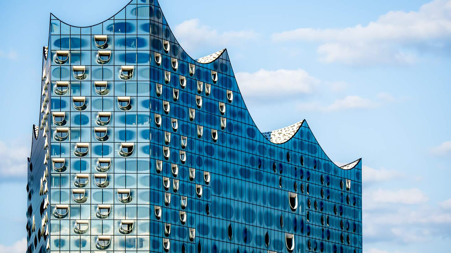 Nahaufnahme der markanten Silhouette der Glasplatte der Hamburger Elbphilharmonie, die ihr glaenzendes und futuristisches Wellendesign vor blauem Himmel zeigt, einem der beliebtesten Wahrzeichen Hamburgs | © Gettyimages.com/Christoph Huber