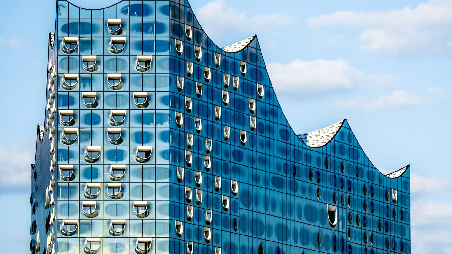 Nahaufnahme der markanten Silhouette der Glasplatte der Hamburger Elbphilharmonie, die ihr glaenzendes und futuristisches Wellendesign vor blauem Himmel zeigt, einem der beliebtesten Wahrzeichen Hamburgs | © Gettyimages.com/Christoph Huber