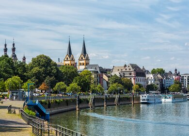 Blick auf die Uferpromenade in Koblenz mit historischen Gebäuden, grünen Bäumen und der markanten Kirche mit zwei Türmen am Rhein unter blauem Himmel. | © Gettyimages.com/	querbeet