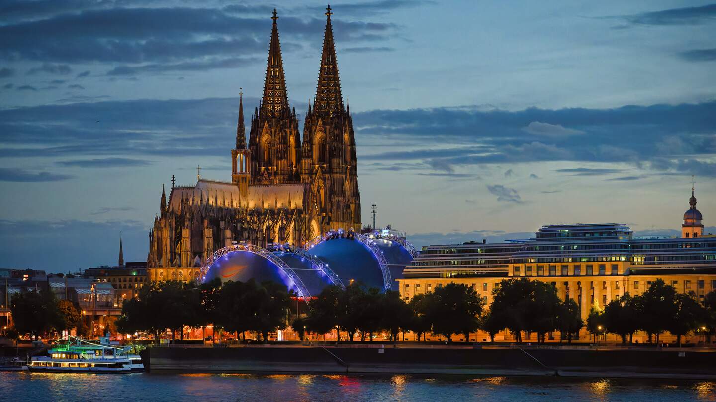 Der beleuchtete Kölner Dom erhebt sich hinter dem blau leuchtenden Musical Dome und dem EASA-Gebäude. Im Vordergrund fließt der Rhein. | © GettyImages.com/Gerd Harder