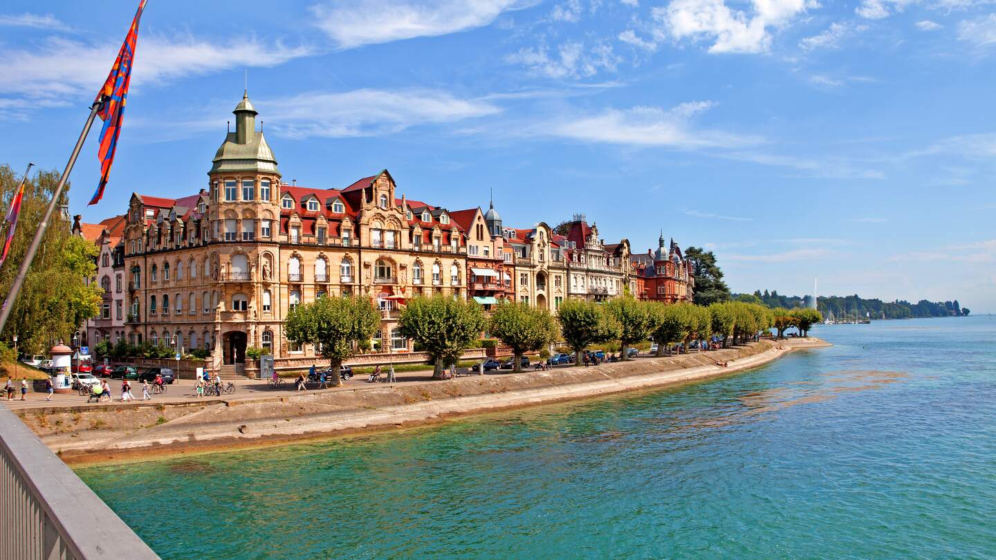 Historische Gebäude mit prachtvollen Fassaden an der Uferpromenade von Konstanz am Bodensee, gesäumt von Bäumen, mit klarem Wasser und blauem Himmel im Hintergrund. | © Gettyimages.com/	lenawurm