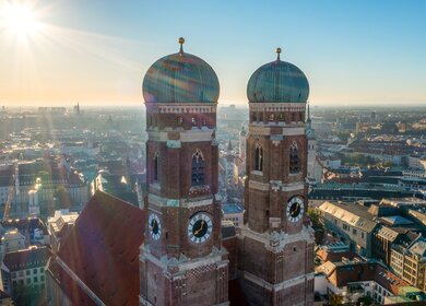 Luftaufnahme der Frauenkirche in München mit ihren markanten Zwiebeltürmen, umgeben von der Stadtlandschaft im warmen Sonnenlicht und klarem Himmel. | © Gettyimages.com/	Luftaufnahme Bayern