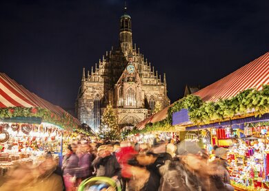 Riesige Menschenmenge, die sich nachts ueber den weltberuehmten Nuernberger Christkindlsmarkt bewegt, vorbei an bunt beleuchteter Weihnachtsdekoration und Imbissstaenden. Nuernbergs Wahrzeichen Frauenkirche ist auf der Rueckseite zu sehen | © Gettyimages.com/Juergen Sack