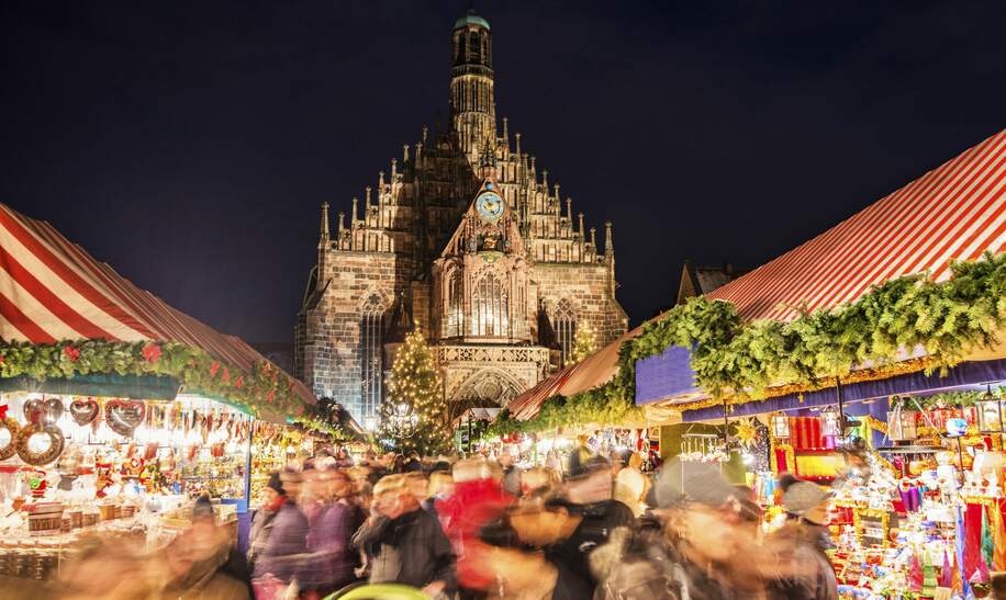 Riesige Menschenmenge, die sich nachts ueber den weltberuehmten Nuernberger Christkindlsmarkt bewegt, vorbei an bunt beleuchteter Weihnachtsdekoration und Imbissstaenden. Nuernbergs Wahrzeichen Frauenkirche ist auf der Rueckseite zu sehen | © Gettyimages.com/Juergen Sack