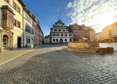 Historischer Marktplatz in Weimar mit Kopfsteinpflaster, Brunnen und dem prächtigen Renaissance-Rathaus, umgeben von farbigen Fassaden unter blauem Himmel mit Sonne. | © Gettyimages.com/Nikada