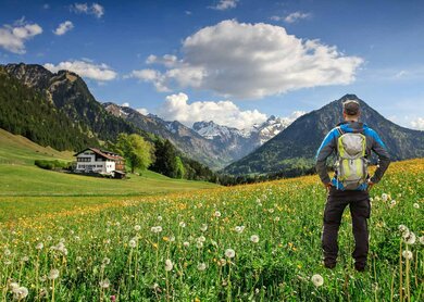 Ein Wanderer mit Rucksack steht auf einer Blumenwiese, umgeben von schneebedeckten Bergen und einem traditionellen Haus in Bayern, den Alpen, Oberstdorf im Allgäu, Deutschland | © Gettyimages.com/Drepicter
