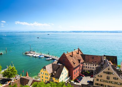 Blick auf den Bodensee von Meersburg aus mit farbenfrohen historischen Häusern im Vordergrund, einem kleinen Hafen mit Booten und dem weiten, blauen See unter klarem Himmel. | © Gettyimages.com/	Michal Krakowiak