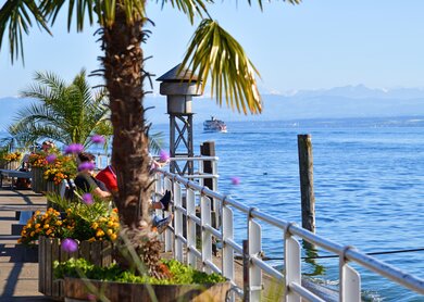 Blühende Blumen und Palmen säumen die Hafenpromenade von Meersburg am Bodensee, mit Blick auf das ruhige Wasser und ein Schiff vor den Alpen im Hintergrund. | © Gettyimages.com/	Peter Allgaier
