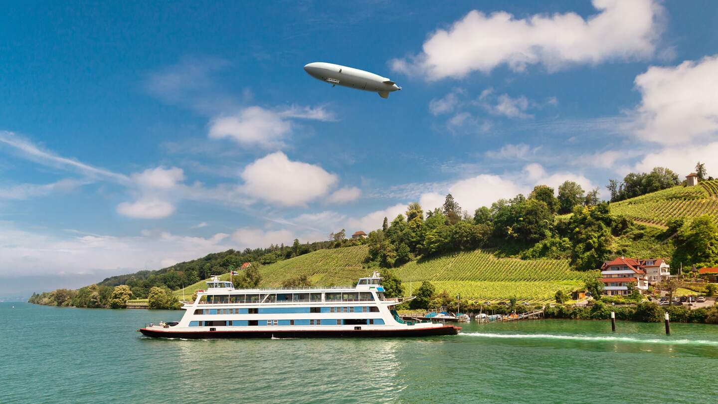 Passagierfähre von Meersburg auf ihrer Route über den Bodensee an einem Sommertsg, mit Weinbergen und einem Zeppelin im Hintergrund | © © Gettyimages.com/olgysha2008