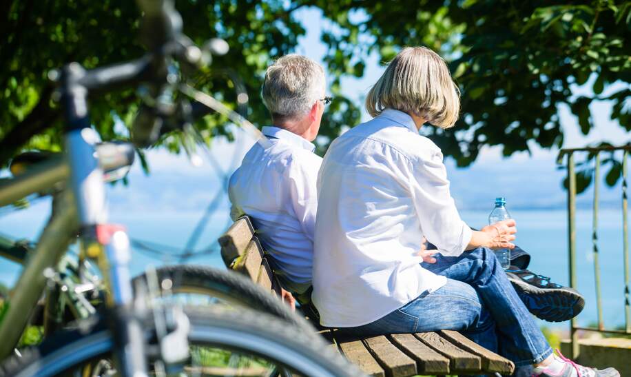 Bestager-Paar genießt bei einer Fahrradtour-Auszeit auf einer Bank die Aussicht auf den Bodensee | © © Gettyimages.com/kzenon