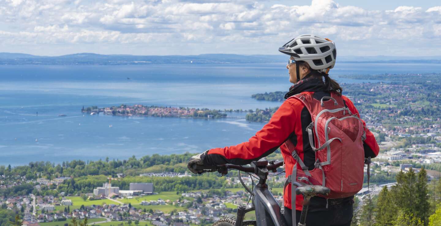 Frau mit Elektro-Mountainbike steht auf einem Berg und schaut hinunter auf das Panorama des Bodensees | © GettyImages.com/Uwe Moser