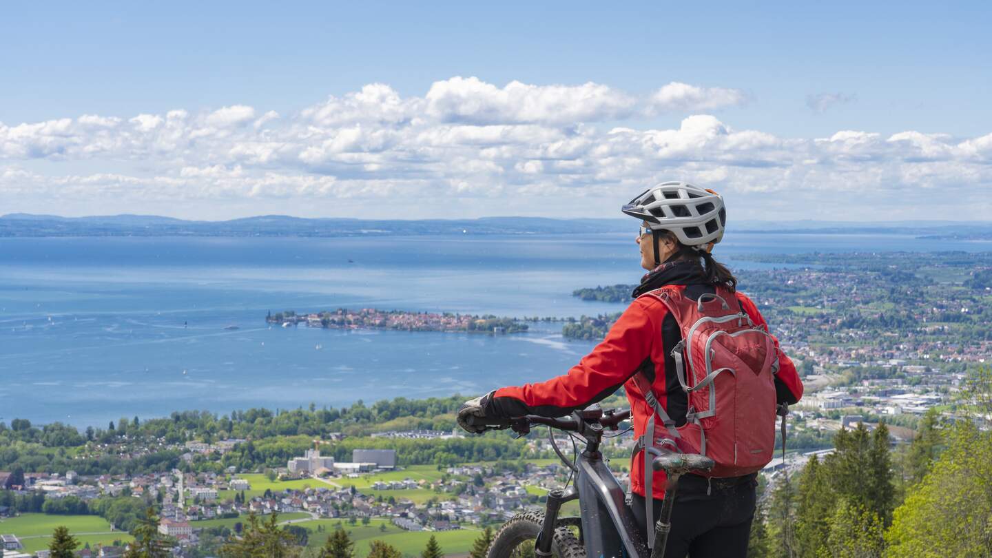Frau mit Elektro-Mountainbike steht auf einem Berg und schaut hinunter auf das Panorama des Bodensees | © GettyImages.com/Uwe Moser