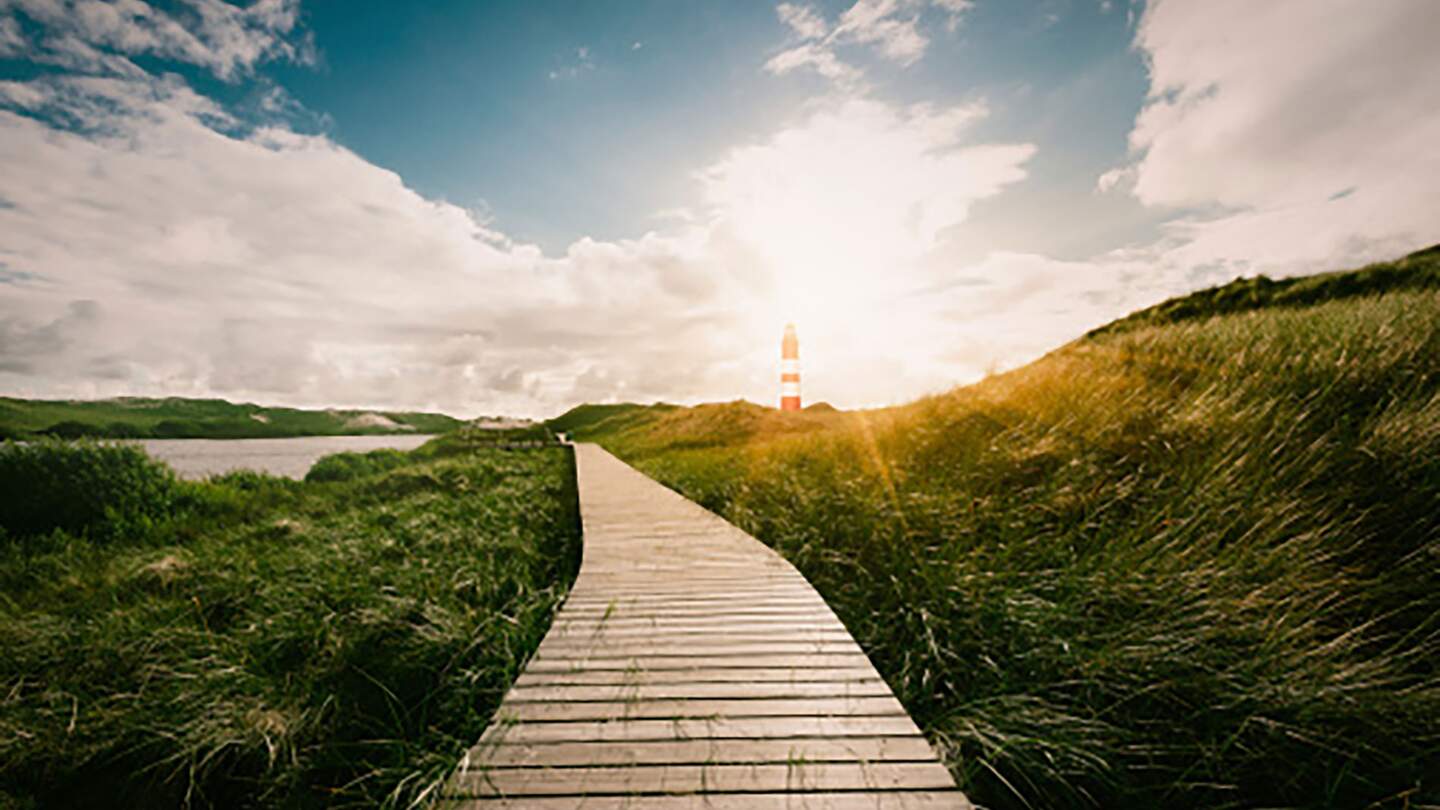 Ein Holzsteg führt bei Sonnenschein durch Wiesen und Wasser zu einem rot weißen Leuchturm | © © Gettyimages.com/Manuel Gutjahr