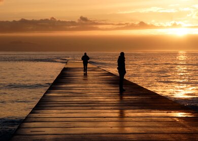 Sonnenuntergang ueber dem Meer mit zwei Sillhouetten, die auf einem steg hervorstechen | © gettyimages.com/ideeone