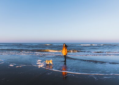 Frau mit langen Haaren und in orangefarbenem Regenmantel und ihr kleiner Hund - Mopsrasse haben eine tolle Zeit am Strand, spielen mit Wellen, laufen und laufen  | © Gettyimages.com/anastasiiashavshyna