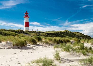  einen rot-weißen Leuchtturm, der auf einer grasbewachsenen Sandduene steht, unter einem blauen Himmel mit verstreuten Wolken. | © gettyimages.com/Oliver Schlünz
