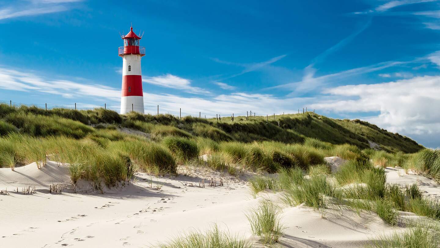  einen rot-weißen Leuchtturm, der auf einer grasbewachsenen Sandduene steht, unter einem blauen Himmel mit verstreuten Wolken. | © gettyimages.com/Oliver Schlünz