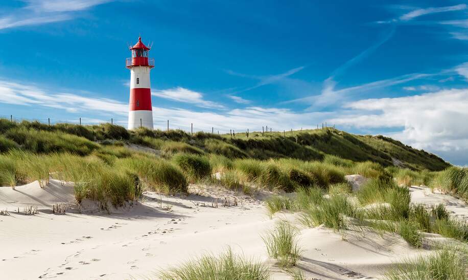  einen rot-weißen Leuchtturm, der auf einer grasbewachsenen Sandduene steht, unter einem blauen Himmel mit verstreuten Wolken. | © gettyimages.com/Oliver Schlünz