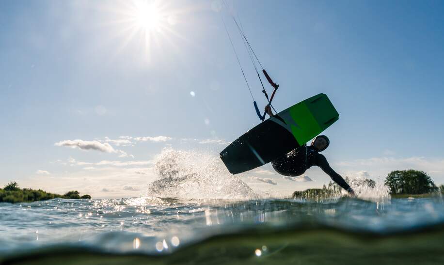 Kitesurfer gleitet mit einer Hand über das Wasser, fotografiert von der Wasseroberfläche aus bei Sonnenschein | © Gettyimages.com/Jens Breuer