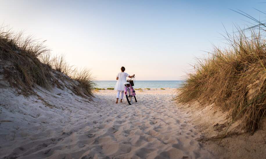 Frau schiebt ein Fahrrad auf einen weissen Sandstrand auf Ruegen mit dem Meer im Hintergrund | © Gettyimages.com/FredFroese