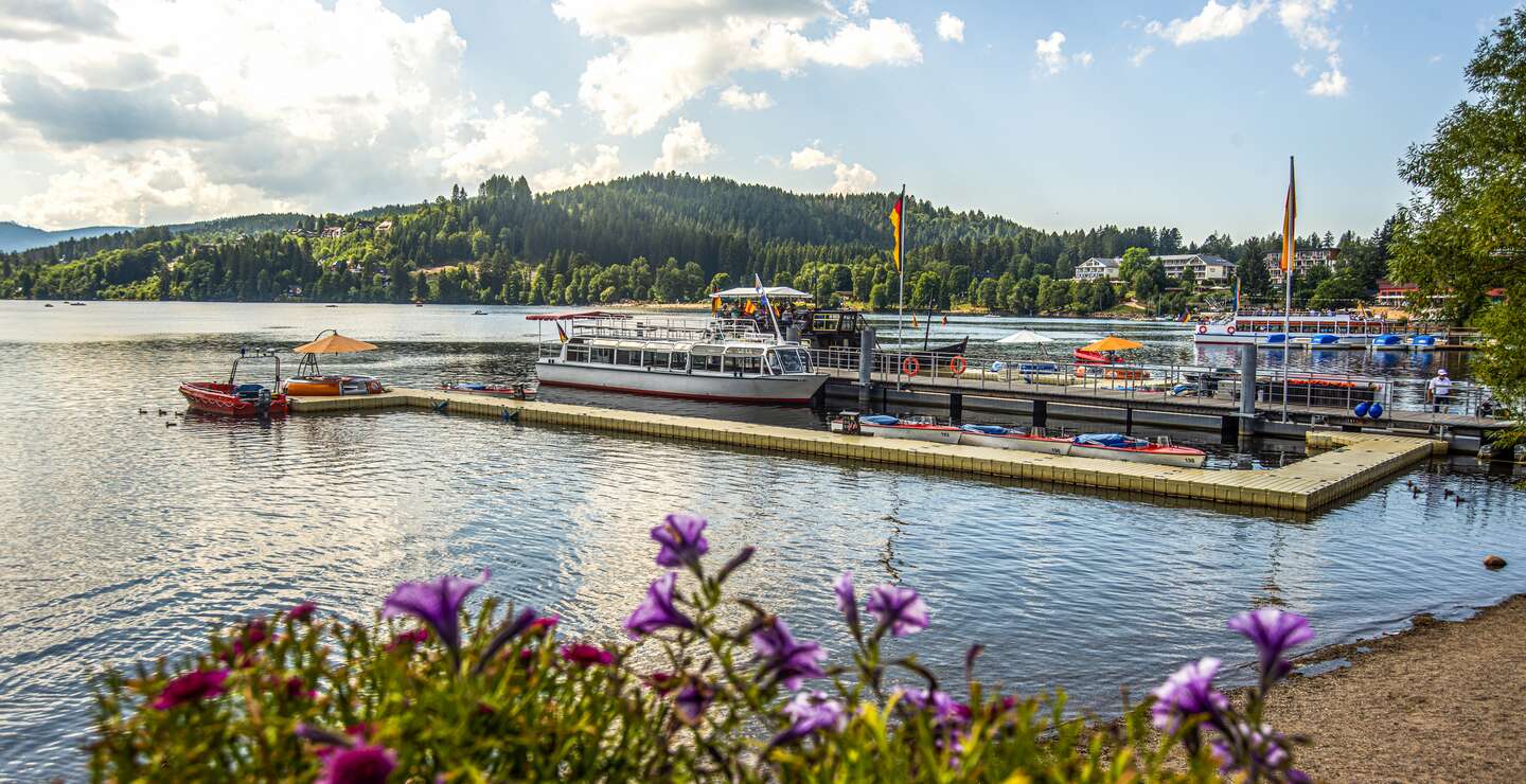 Blick auf einen Bootsanleger mit Booten auf dem Titisee bei Sonnenschein und leichten Wolken | © Gettyimages.com/Wirestock