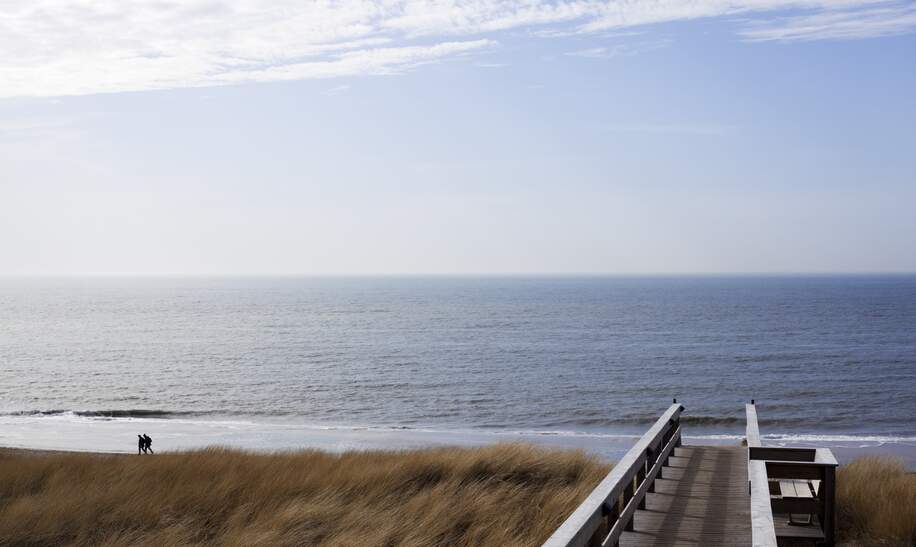 Ein Holzsteg fürht zu einer Treppe mit dessen Hilfe man sich auf den Strand begeben kann. Links im Bild laufen zwei Personen. Der ganze Hintergund wird von dem Himmel und dem Meer eingenommen.  | © Gettyimages.com/alexhagmann