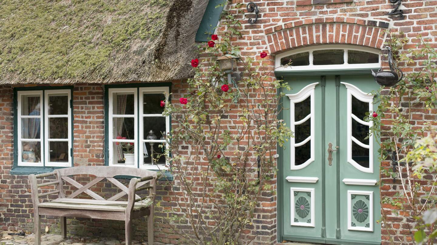 Fassade eines typischen traditionellen Zuhauses auf der Insel Sylt, Deutschland. Die gruene Tür wird von roten Rosen umramt. Vor dem Haus steht eine Bank | © Gettyimages.com/relaxfoto.de