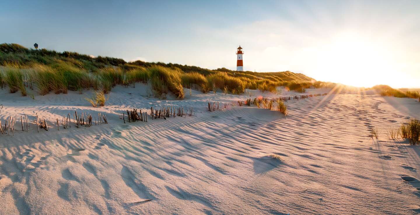 Ein rot weißer Leuchtturm in den Dünen, die Sonne geht unter und taucht die Grashalme auf den Dünen in goldfarbenes Licht | © Gettyimages.com/mthaler