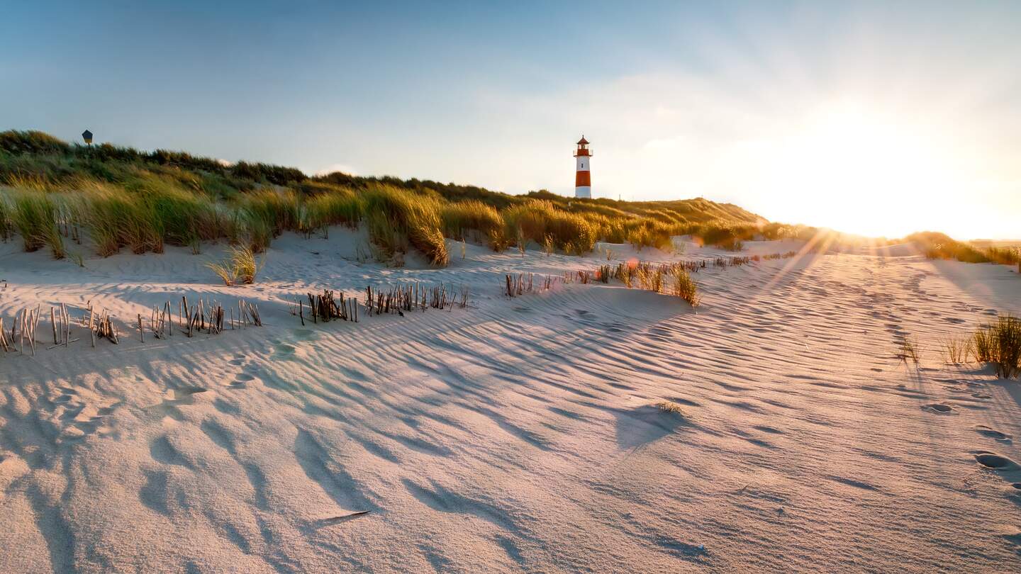 Ein rot weisser Leuchtturm in den Duenen, die Sonne geht unter und taucht die Grashalme auf den Duenen in goldfarbenes Licht | © Gettyimages.com/mthaler