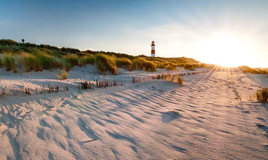 Ein rot weisser Leuchtturm in den Duenen, die Sonne geht unter und taucht die Grashalme auf den Duenen in goldfarbenes Licht | © Gettyimages.com/mthaler