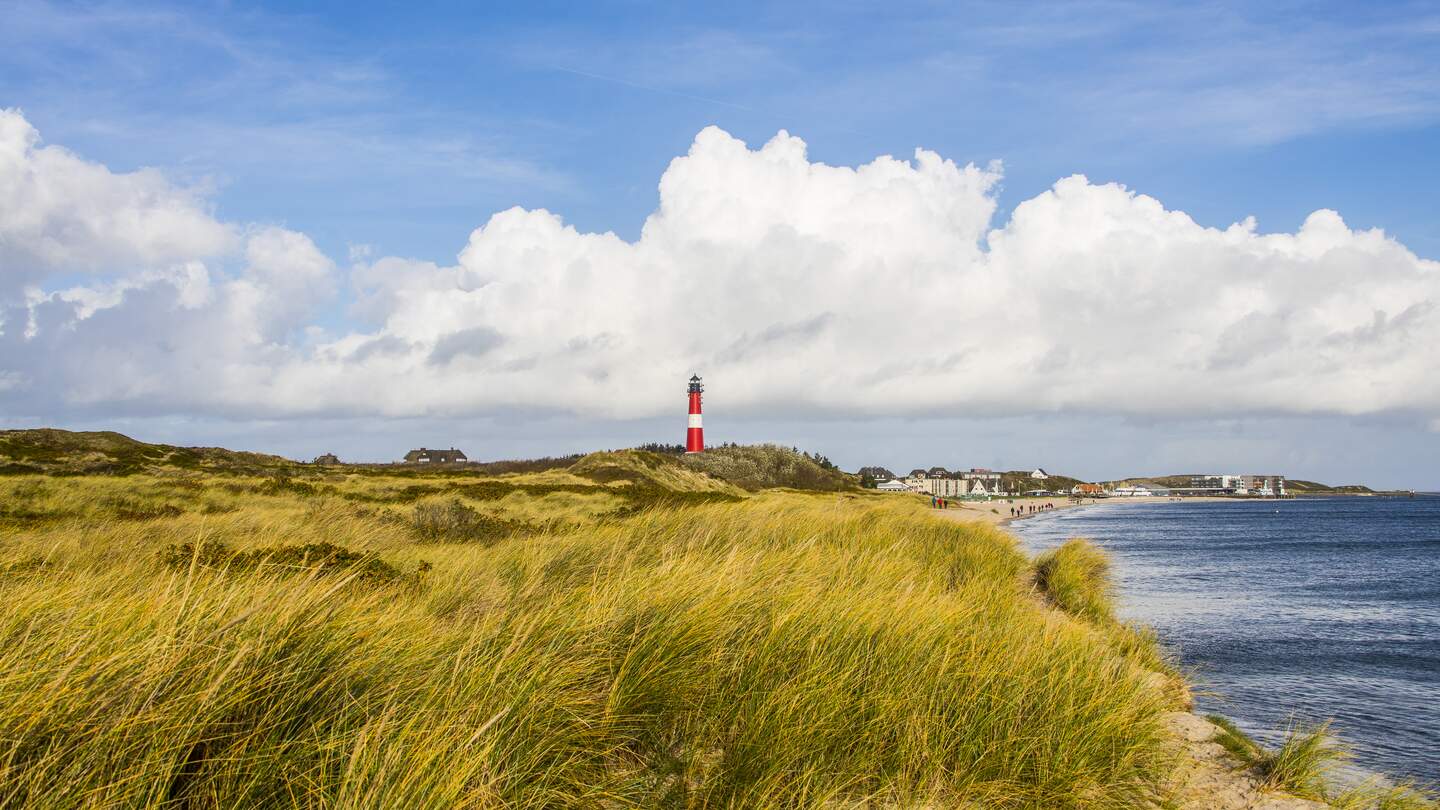 Der rot, weiße Leuchtturm ragt in den Dünen empor. Im Hintergrund sieht man den Strand mit Menschen und ein paar Häuser: Rechts ist das Meer. | © Gettyimages/marcstephan