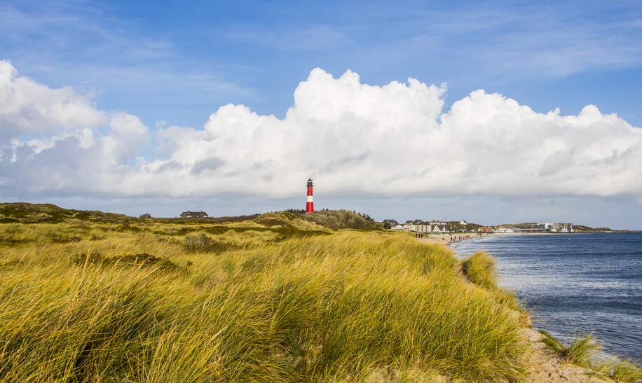 Der rot, weiße Leuchtturm ragt in den Dünen empor. Im Hintergrund sieht man den Strand mit Menschen und ein paar Häuser: Rechts ist das Meer. | © Gettyimages/marcstephan