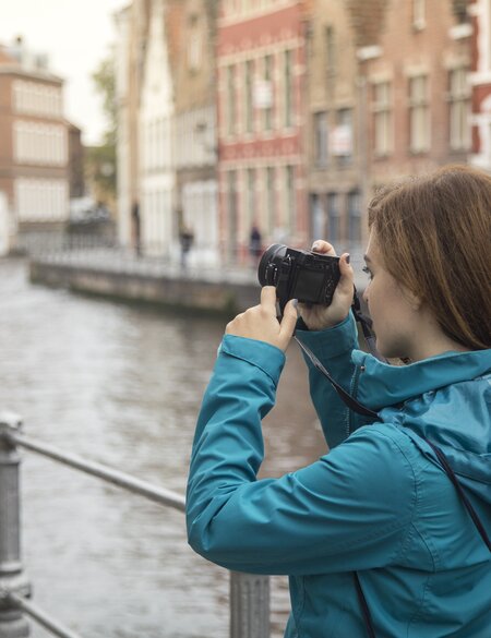 Weibliche Touristin fotografiert in Brügge an einem Kanal, in dem ein Boot fährt. | © Gettyimages.com/GoodLifeStudio