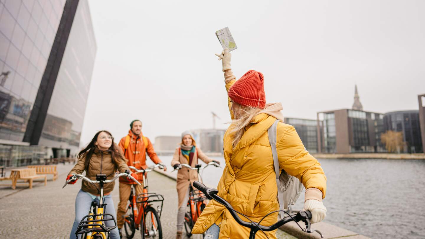 Gruppe Touristen mit dicken Jacken und Mützen auf Fahrrädern am Hafen in Kopenhagen, Dänemark, vor der Det Kongelige Bibliothek | © GettyImages.com/AleksandarNakic