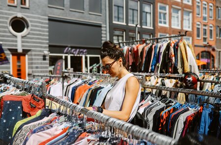 Frau mit Sonnenbrille und ärmelloser Bluse schaut durch die Kleiderständer eines Flohmarktes in Amsterdam | © Gettyimages.com/lechatnoir